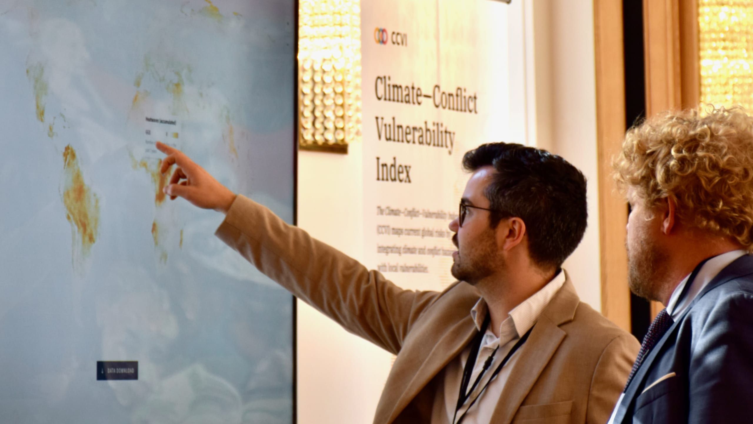 Analyst pointing at a projected CCVI world map beside a Climate—Conflict—Vulnerability Index poster